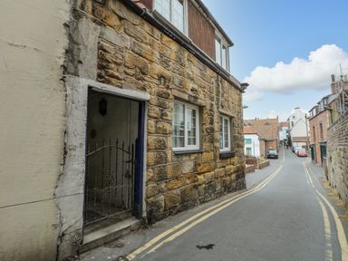 A narrow street with stone and brick buildings and cars parked along the side at April Cottage in Whitby