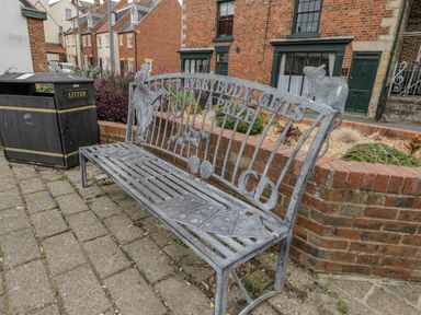 A metal bench with playing card and rat designs next to a litter bin on a paved sidewalk near brick buildings at April Cottage in Whitby