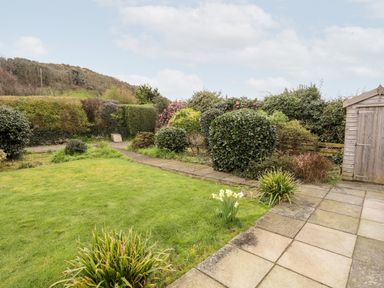 A garden with grass lawn and bushes next to a stone patio and a wooden shed at Bro Mebyd in Criccieth