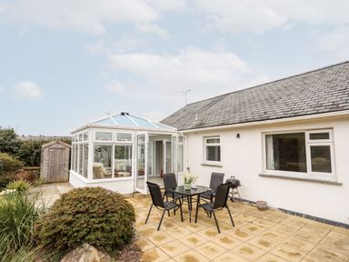 An outdoor patio with a glass table and four chairs next to a white house and a conservatory at Bro Mebyd in Criccieth