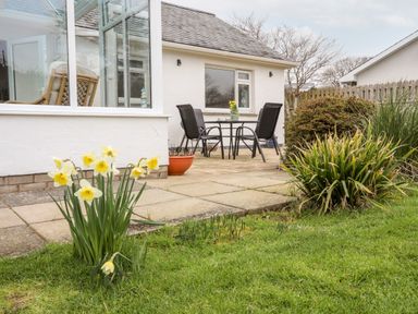 A patio with a table and four chairs outside a white house with daffodils and bushes in the garden at Bro Mebyd in Criccieth