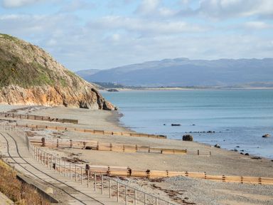 A pebble beach with wooden groynes and a concrete path with railings next to a rocky hill and calm sea at Bro Mebyd in Criccieth