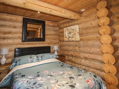 A bedroom with a bed with floral bedding two bedside tables with lamps a wall mirror and artwork on wooden log walls at Brynallt Country Park Frankton near Ellesmere