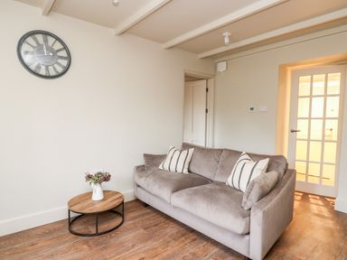 A living room with a gray sofa two striped pillows a round wooden coffee table with a vase of flowers and a wall clock at Llygad Y Dydd in Bow Street Wales