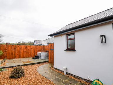 An outdoor patio area with a hot tub on a stone platform surrounded by a wooden fence and a gravel pathway at Llygad Y Dydd in Bow Street Wales