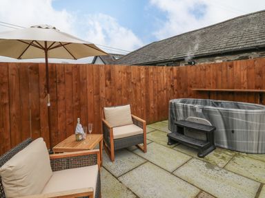 An outdoor patio with two cushioned chairs a wooden table a large umbrella and a hot tub at Llygad Y Dydd in Bow Street Wales