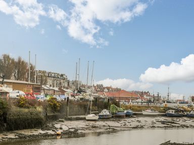 Boats docked on muddy shore near stone retaining wall and buildings under blue sky at Estuary Walk in Whitby
