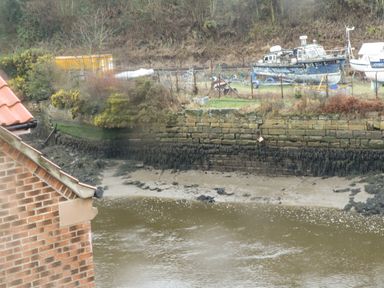 A muddy riverbank with boats on land and a brick building on the left at Estuary Walk in Whitby