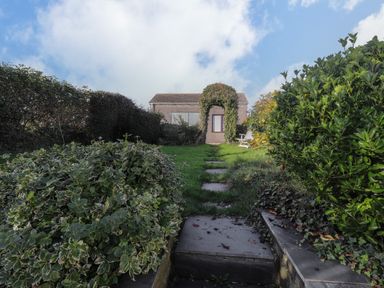 A stone path through a garden with bushes leading to a small building with a door covered by an arch of climbing plants at Hillcrest Oxwich near Port Eynon