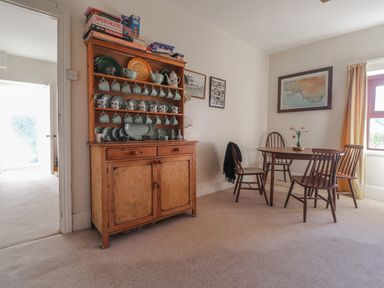 A dining area with a wooden cupboard holding dishes and board games and a wooden table with four chairs and a vase with flowers at Hillcrest in Oxwich near Port Eynon