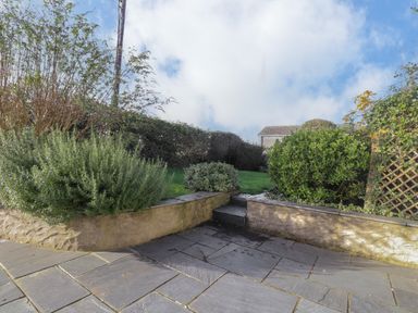 An outdoor patio with stone tiles and a small step leading to a grassy area with bushes and hedges at Hillcrest in Oxwich near Port Eynon