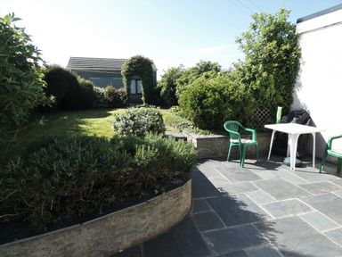 A garden with bushes lawn and a small building viewed from a stone patio with plastic chairs and a white table at Hillcrest in Oxwich near Port Eynon