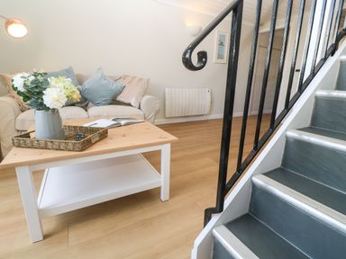 A living room with a beige sofa, blue and beige cushions, a wooden coffee table with a flower vase and a book, next to stairs with a black handrail at 58 High Street in Falmouth