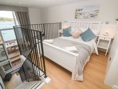 A bedroom with a white bed and bedside tables next to a stairway and large window overlooking water at 58 High Street in Falmouth