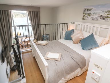 A bedroom with a white bed and pillows next to a railing overlooking a balcony with a dining table at 58 High Street in Falmouth