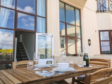 A wooden outdoor dining table set with bowls plates glasses and a champagne bottle on a balcony with large windows at 58 High Street in Falmouth