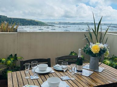A balcony with a wooden table set with bowls glasses a bottle and flowers overlooking a harbor with boats at 58 High Street in Falmouth