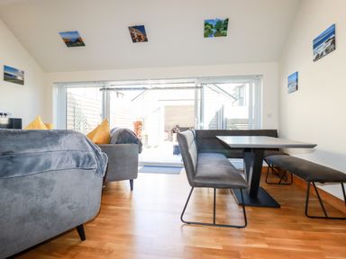 A dining area with a rectangular table and bench seating next to a living area with grey sofas and yellow cushions at Clonmore in Bodmin