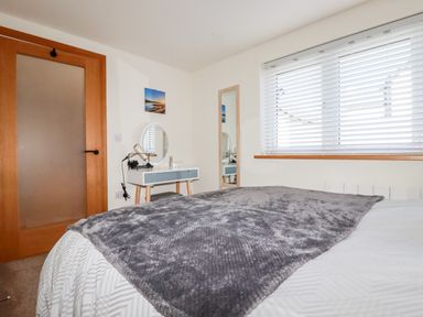 A bedroom with a bed covered with a gray blanket a wooden door a vanity table with a lamp and round mirror a tall mirror on the wall and a window with blinds at Clonmore in Bodmin