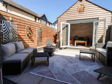 An outdoor patio with cushioned seating, metal wall decor, a central fire pit, and open doors leading to an indoor room at Clonmore in Bodmin