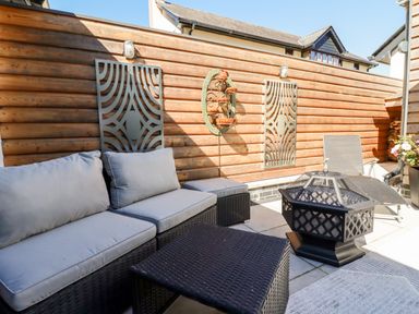 An outdoor patio with a wicker sofa with cushions a coffee table a fire pit a lounge chair and wood panel fencing with decorative metal wall art at Clonmore in Bodmin
