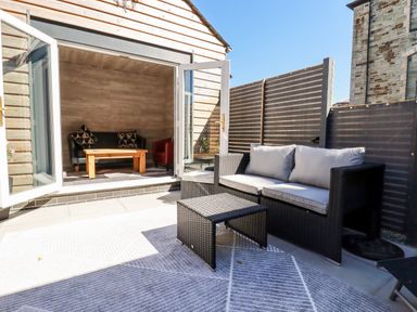 An outdoor patio area with a wicker sofa and table next to open double doors leading to a small room with a black sofa red armchair and wooden coffee table at Clonmore in Bodmin