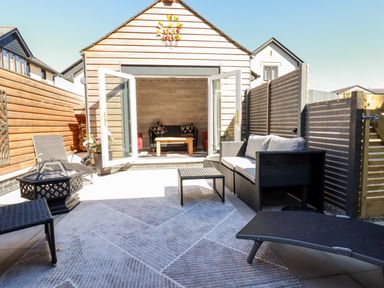 An outdoor patio area with wicker seating and a table near a wooden building with open double doors at Clonmore in Bodmin