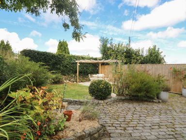 A garden with plants a stone patio and a wooden pergola with white outdoor furniture at Trelash Cottage in Trelash near Boscastle
