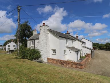 A white stone cottage with chimneys next to a grassy area and a dirt road at Trelash Cottage in Trelash near Boscastle
