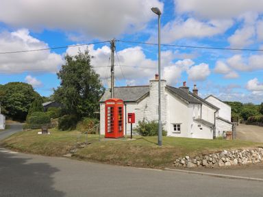 A white cottage with chimneys next to a red telephone box and a red post box on a grassy area with a lamp post and a stone wall at Trelash Cottage in Trelash near Boscastle