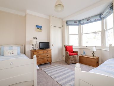 A bedroom with two single beds a wooden chest of drawers a wicker chair with red cushion and a wooden chest under bay windows at Driftwood in Fowey