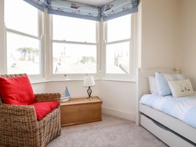 A corner bedroom with a wicker chair with red cushions a wooden chest a lamp a small sailboat model and a white bed with blue and white bedding at Driftwood in Fowey