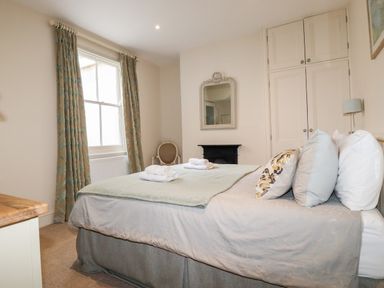 A bedroom with a bed pillows folded towels an armchair a mirror a fireplace built in cupboard and window with curtains at Driftwood in Fowey
