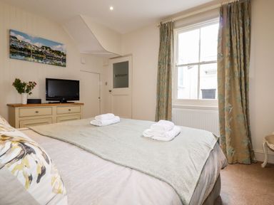 A bedroom with a bed, folded towels, a dresser with a TV and flowers, a window with curtains, and a door labeled bathroom at Driftwood in Fowey