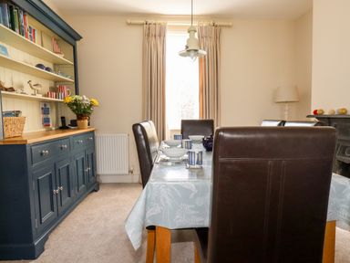 A dining room with a table set with bowls cups and a teapot brown chairs a blue cabinet with shelves and a window with curtains at Driftwood in Fowey
