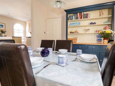 A dining room with a table set with bowls mugs and cutlery and a blue cabinet with books and decorations at Driftwood in Fowey