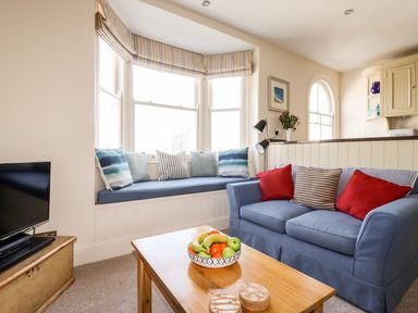 A sitting area with a blue sofa and striped cushions next to a window seat with cushions and a wooden coffee table with a bowl of fruit at Driftwood in Fowey