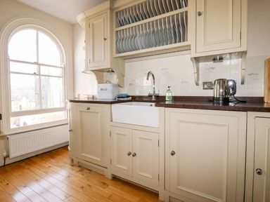 A kitchen with cream cabinets a white farmhouse sink a kettle and plates stored above at Driftwood in Fowey