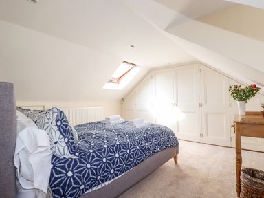 A bedroom with a bed covered by blue patterned bedding next to a skylight window and built in white closets with a wooden table and flowers at Driftwood in Fowey