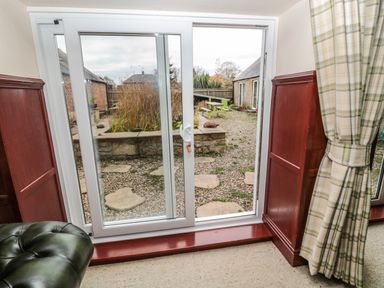 Sliding glass door with wooden paneling and checked curtain looking out to a garden with stepping stones and green picnic tables at The Granary in Longhirst near Morpeth