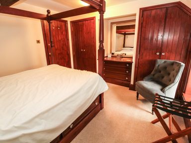 A bedroom with a four-poster bed wooden wardrobes a dresser with a mirror a gray armchair and a luggage rack at The Granary in Longhirst near Morpeth