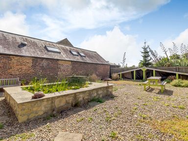 An outdoor area with a raised stone pond surrounded by plants gravel ground a green picnic table and a brick building with skylights at The Granary in Longhirst near Morpeth