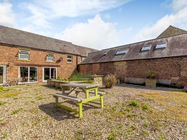 An outdoor area with a wooden picnic table and gravel ground next to brick buildings at The Granary in Longhirst near Morpeth