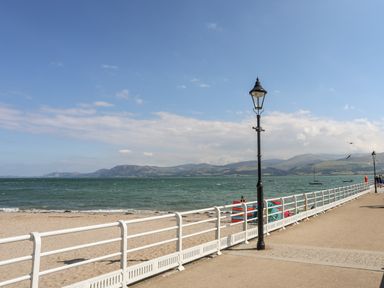 A seaside promenade with white railings and lampposts along a sandy beach by the sea with hills in the background at Above Deck in Beaumaris