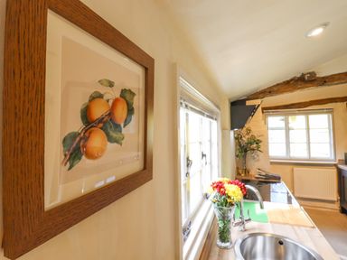 A kitchen with a framed fruit painting on the wall flowers in a vase on the counter and windows at The Abbey Cottage in Windsor Green