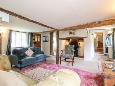 A living room with sofas a patterned armchair a fireplace wooden beams and windows with curtains at The Abbey Cottage in Windsor Green