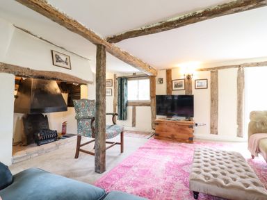 A living room with exposed wooden beams a fireplace a patterned armchair a television on a wooden chest a pink rug and a tufted ottoman at The Abbey Cottage in Windsor Green