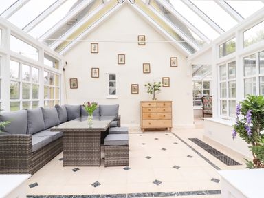 A conservatory with a grey cushioned seating area a glass table a wooden dresser and framed pictures on the wall at The Abbey Cottage in Windsor Green