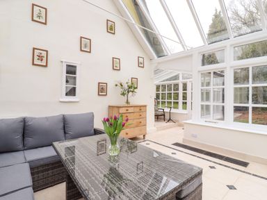 A conservatory with a glass table holding a vase of purple tulips a gray cushioned sofa a wooden chest of drawers and framed pictures on the wall at The Abbey Cottage in Windsor Green