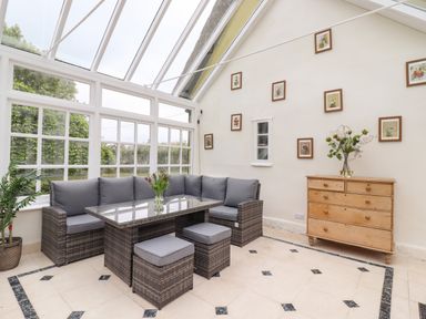 A conservatory with a wicker sofa and stools around a glass table and a wooden dresser with flowers at The Abbey Cottage in Windsor Green